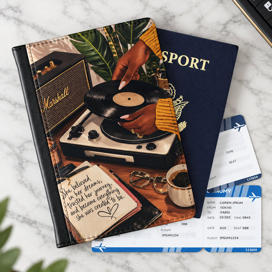 A notebook or planner with a travel-themed cover design lies on a wooden tabletop, surrounded by travel props including a passport, boarding pass, ticket, mug, glasses, and a key fob. The cover shows a person placing a vinyl record on a record player with a Marshall speaker, palm leaves, and a quote notebook.