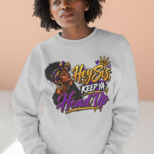 A woman wearing a light gray crewneck sweatshirt with a colorful graphic print that reads 'Hey Stfu, Keep Ya Head Up' in gold, purple, and black lettering, paired with blue jeans, set against a warm peach-colored wall background.