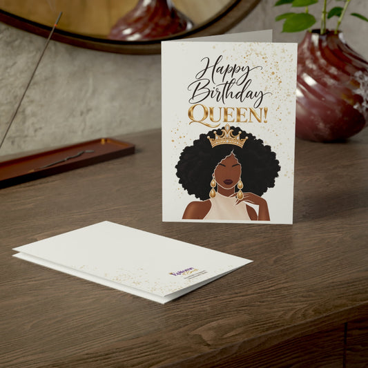 A birthday greeting card and matching envelope displayed on a wooden table. The card features the text 'Happy Birthday QUEEN!' in black and gold script with an illustration of a Black woman wearing a gold crown and earrings, set against a white background with subtle gold speckles.