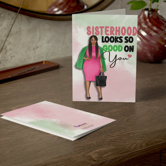 A greeting card and matching envelope displayed on a wooden table. The standing card features a pastel pink and mint watercolor background with an illustration of a woman in a pink dress and green jacket, alongside the text 'SISTERHOOD LOOKS SO GOOD ON You'.