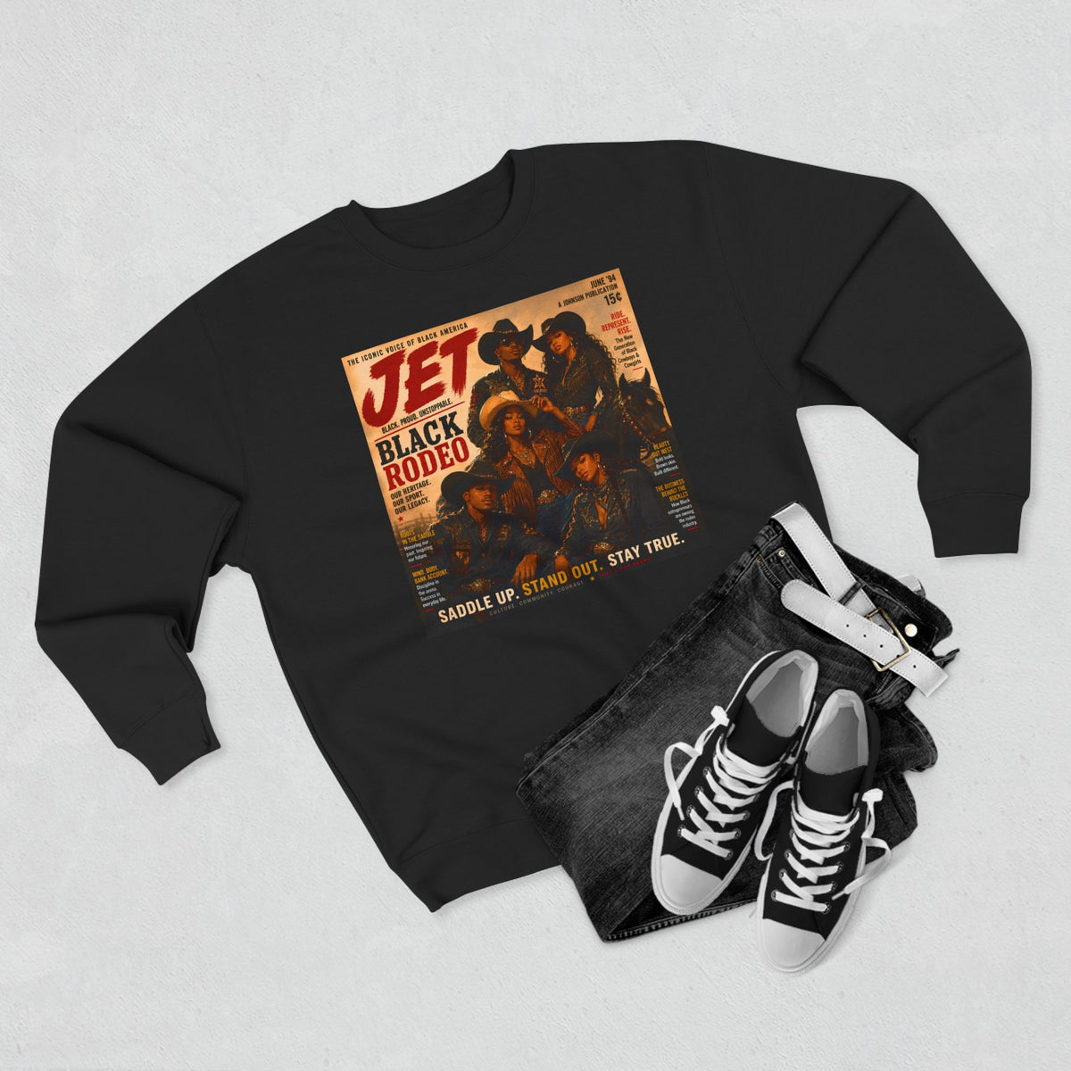A black crewneck sweatshirt featuring a vintage-style 'Jet Black Rodeo' graphic print, styled in a flat lay with dark denim jeans, a white belt, and black-and-white sneakers on a light gray textured background.
