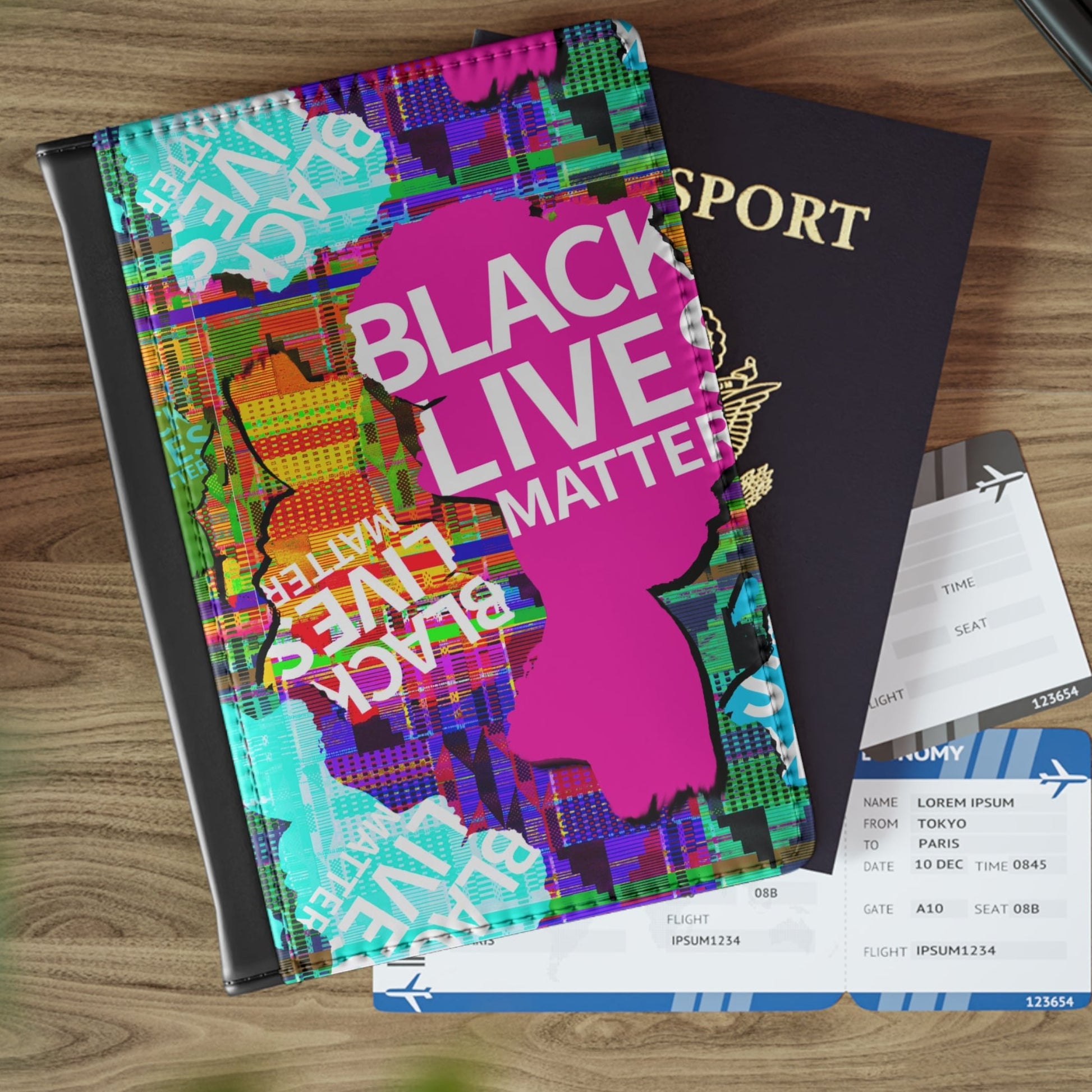 A black binder or folder with a colorful 'Black Lives Matter' graphic cover is arranged on a wooden tabletop next to a passport, car key fob, and boarding pass.