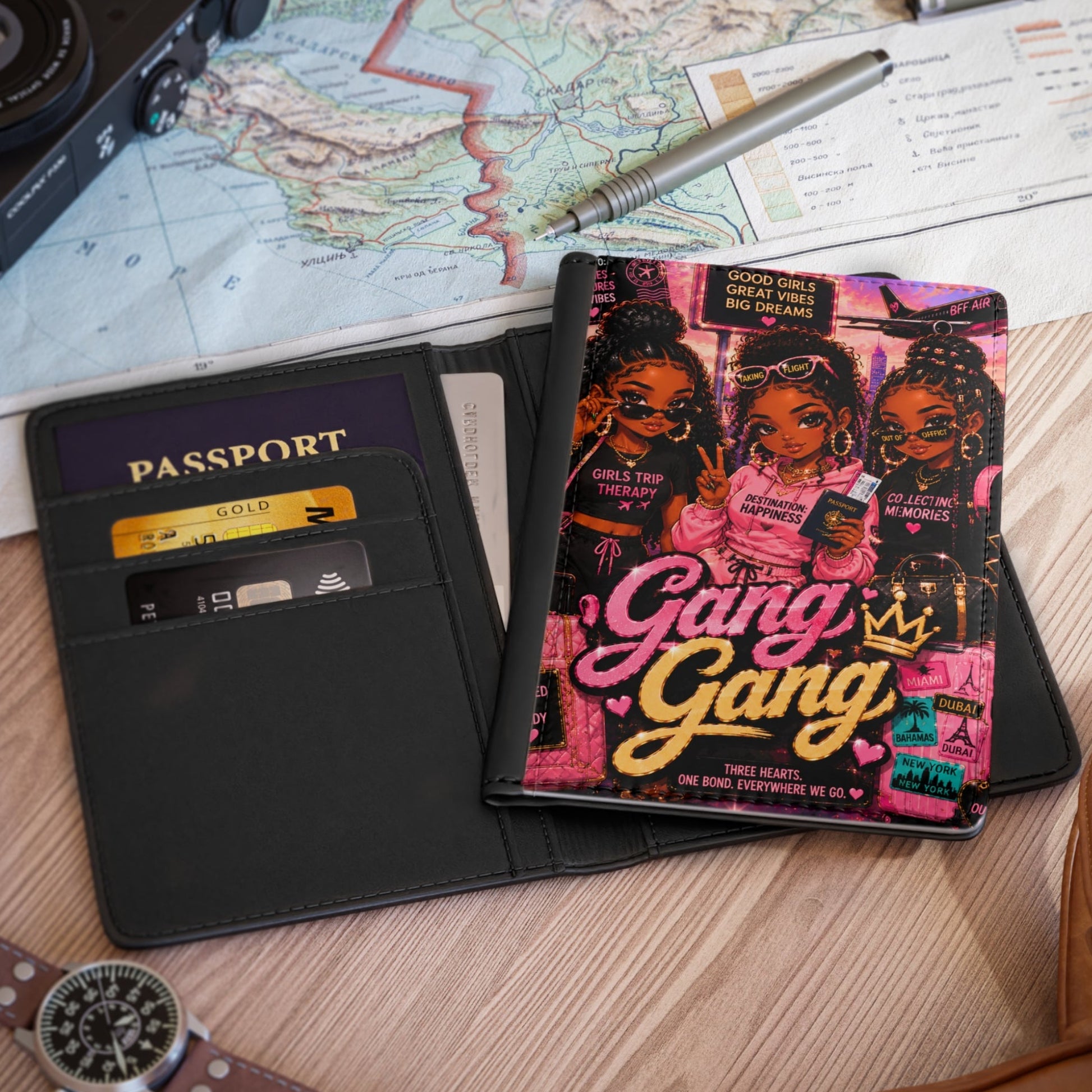 A black leather passport holder wallet shown open on a wooden desk, with a colorful 'Gang Gang' art card or booklet inserted. Surrounding props include a map, camera, pen, passport, watch, and part of a brown leather bag.