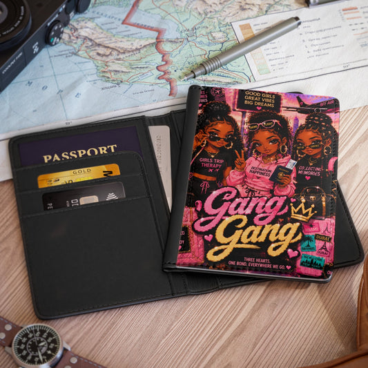 A black leather passport holder wallet shown open on a wooden desk, with a colorful 'Gang Gang' art card or booklet inserted. Surrounding props include a map, camera, pen, passport, watch, and part of a brown leather bag.