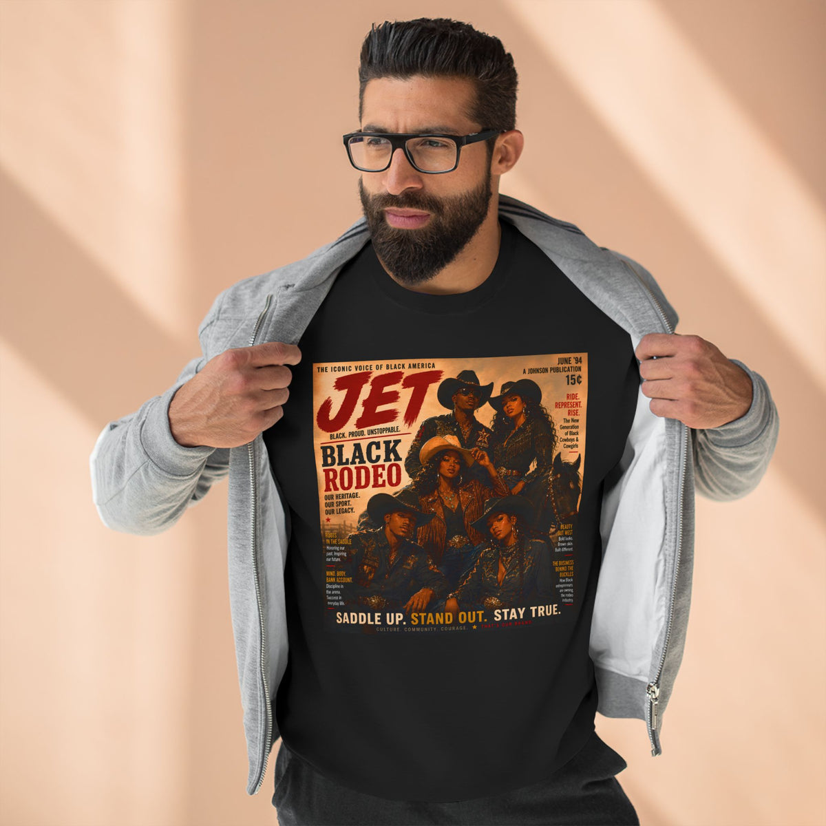 A man wearing a black graphic t-shirt with a vintage-style 'Jet Black Rodeo' print, layered under an open light gray zip-up hoodie. He is wearing glasses and posing against a warm beige background with soft diagonal shadows.