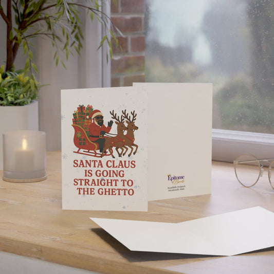 A greeting card displayed on a wooden windowsill with a candle, plant, and eyeglasses nearby. The card shows an illustration of Santa Claus in his sleigh and the text 'SANTA CLAUS IS GOING STRAIGHT TO THE GHETTO'.