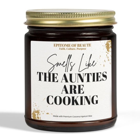 A dark amber glass jar candle with a gold metal lid and a white label that reads 'Smells Like THE AUNTIES ARE COOKING' on a clean white background.