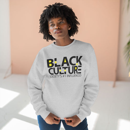 A woman with short curly black hair wearing a light gray crewneck sweatshirt with 'BLACK CULTURE SOCIETY'S #1 INFLUENCED' printed in black and yellow on the front, paired with blue jeans, standing in a brightly lit room with a peach-colored wall.
