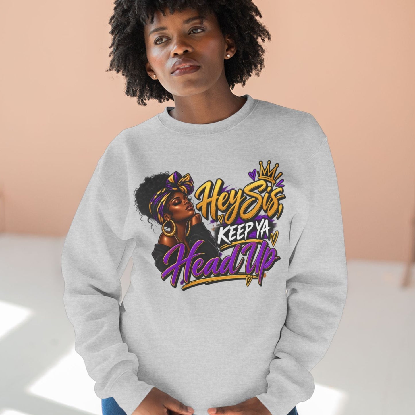 A woman wearing a light gray crewneck sweatshirt with a colorful graphic print that reads 'Hey Stfu, Keep Ya Head Up' in gold, purple, and black lettering, paired with blue jeans, set against a warm peach-colored wall background.