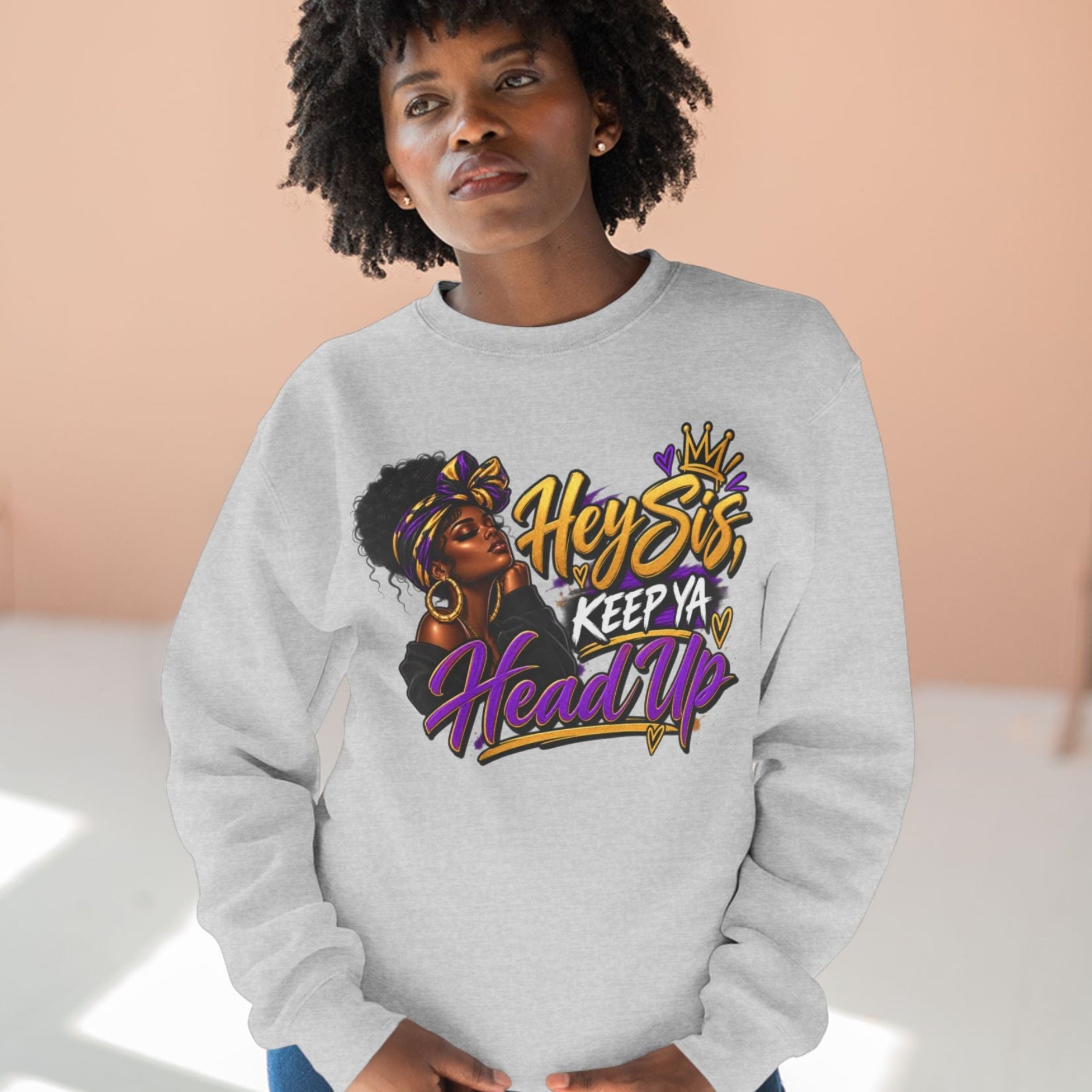A woman wearing a light gray crewneck sweatshirt with a colorful graphic print that reads 'Hey Stfu, Keep Ya Head Up' in gold, purple, and black lettering, paired with blue jeans, set against a warm peach-colored wall background.