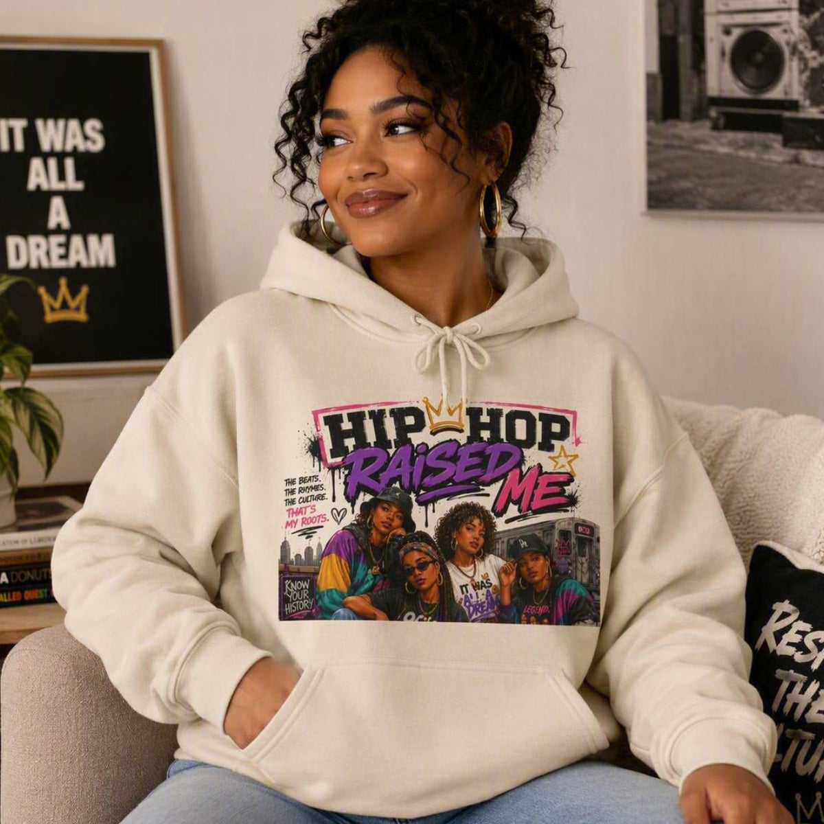 A woman wearing a cream-colored hoodie with a colorful 'Hip-Hop Raised Me' graphic print, sitting on a couch in a living room setting.