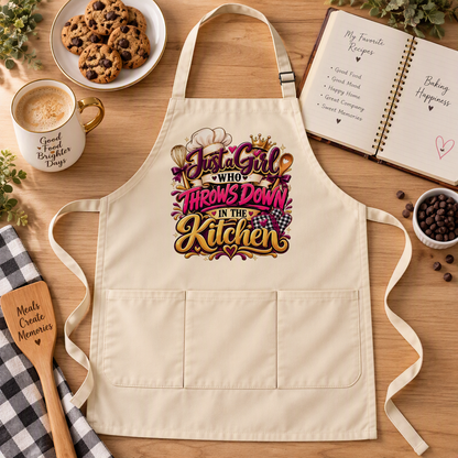 A cream-colored kitchen apron with a colorful graphic reading 'Just a Girl Who Throws Down in the Kitchen' in pink, purple, and gold lettering. The apron is displayed in a styled flat lay on a wooden table surrounded by baking props, including a coffee mug, chocolate chip cookies, an open recipe book, a wooden spoon, and coffee beans.