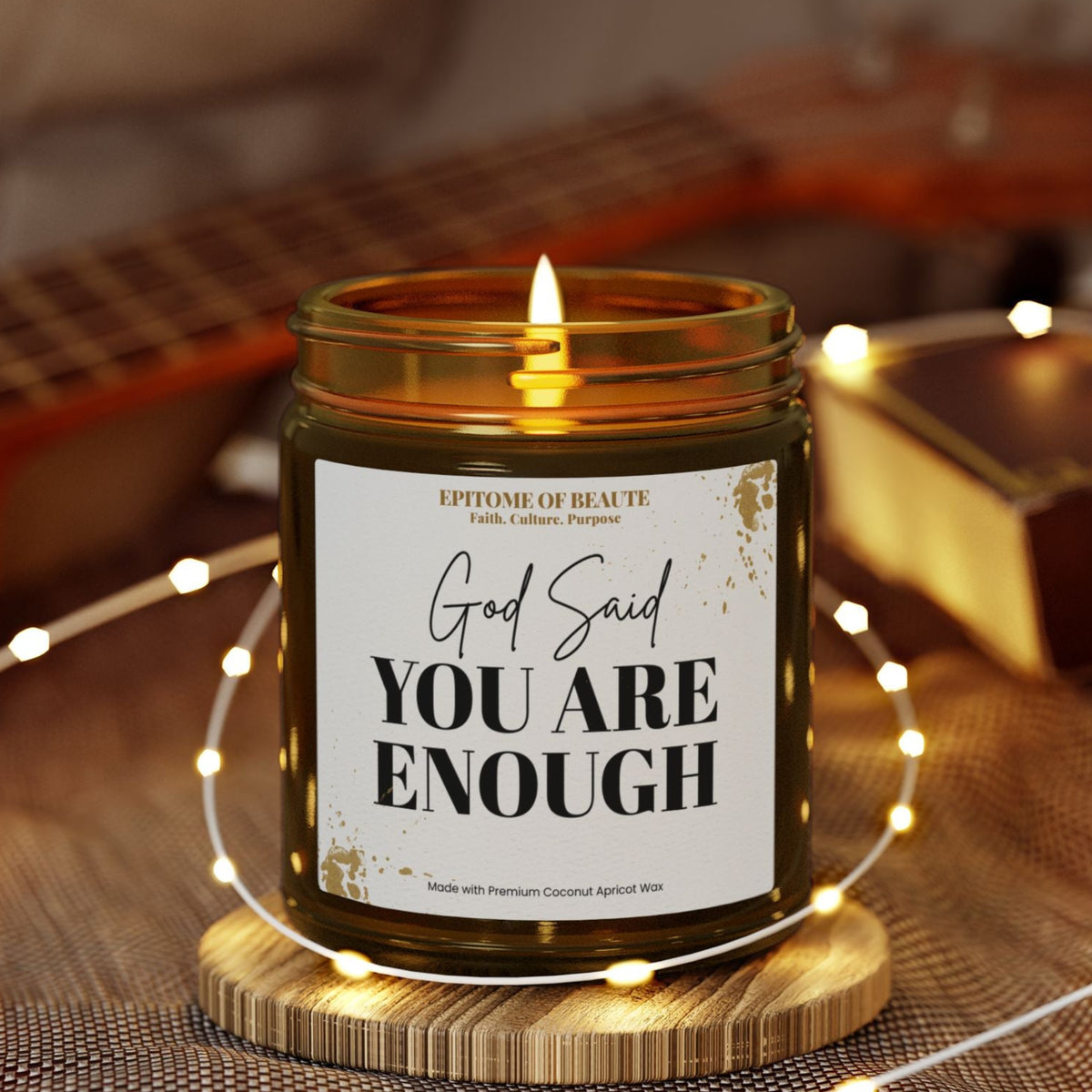 An amber glass jar candle with a white label reading 'God Said YOU ARE ENOUGH' sits lit on a wooden coaster, surrounded by warm fairy lights and cozy home decor in a softly blurred setting.