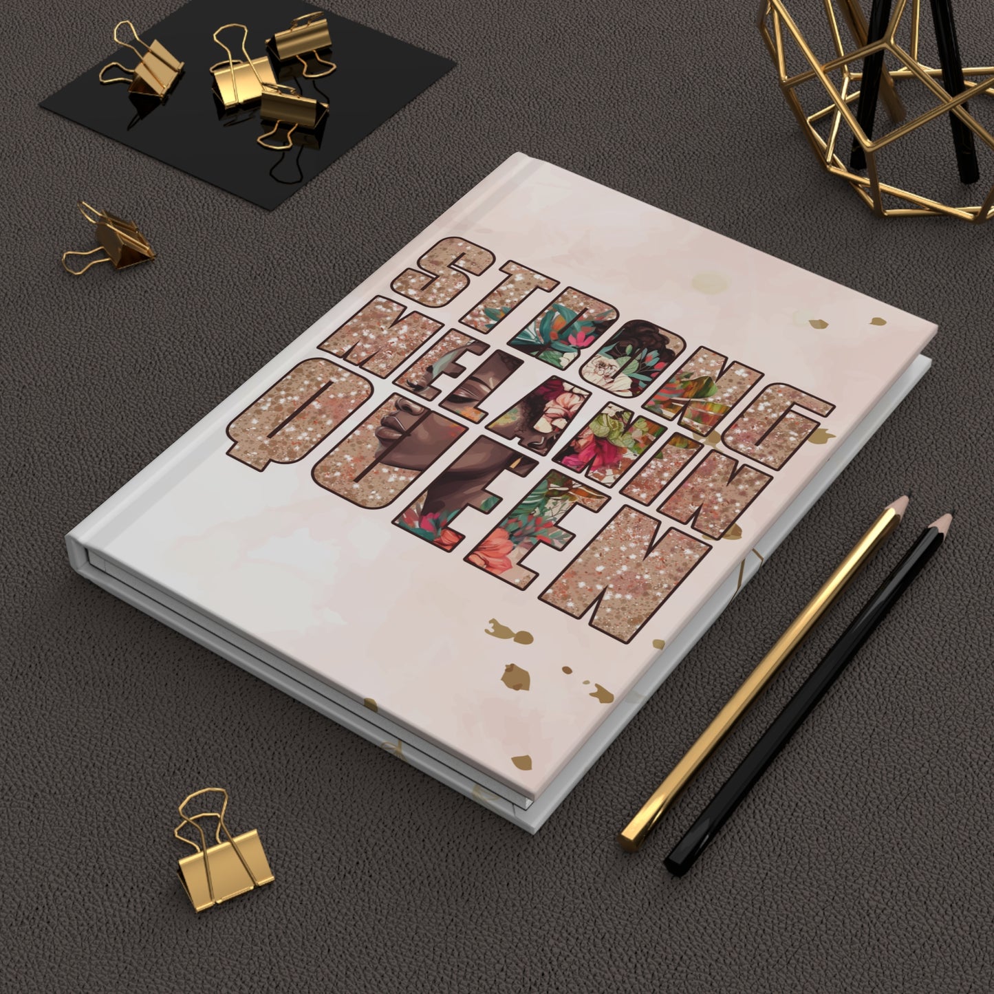 A hardcover notebook with a light pink cover and the words ''STRONG MELANIN QUEEN' in large floral-patterned letters, styled on a dark textured surface with gold binder clips, pencils, and a geometric gold object.