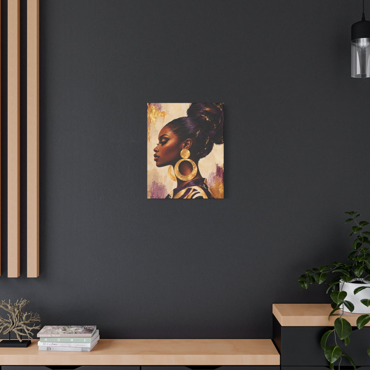 A square wall art print showing a profile portrait of a Black woman with gold hoop earrings, displayed on a dark charcoal wall above a light wood console with a plant and decorative objects.