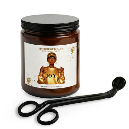 An amber glass jar candle with a black lid and a white label reading 'EPITOME DE BEAUTÉ' and 'JOY', shown with black metal tongs on a clean white background.
