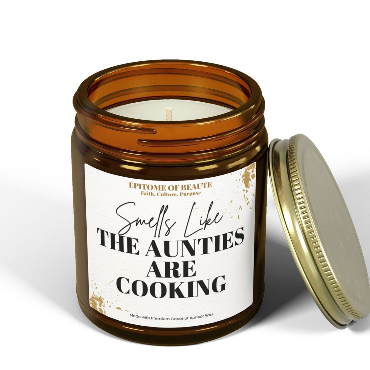 An amber glass jar candle with a white label reading 'Smells Like The Aunties are Cooking,' shown with its gold-toned metal lid leaning against it on a white studio background.