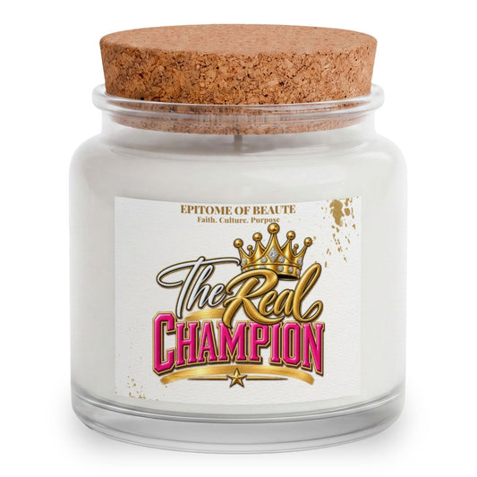 A white wax candle in a clear glass jar with a natural cork lid, featuring a white label with the text 'The Real Champion' in gold and pink lettering with a crown graphic. The brand 'Epitome of Beaute' is printed at the top of the label.