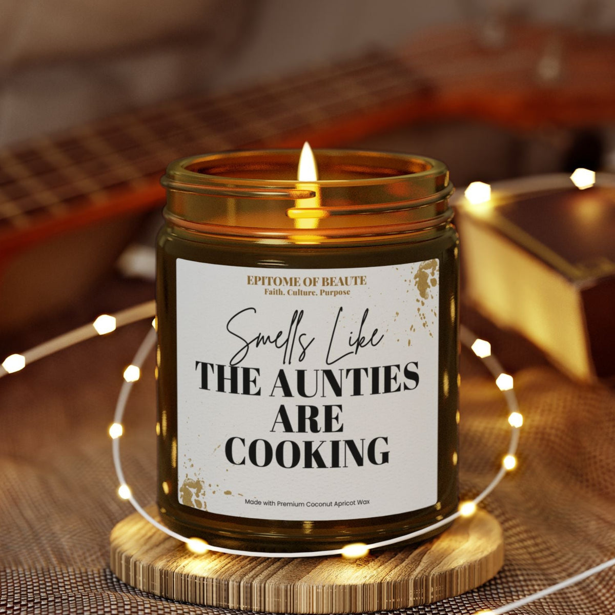 An amber glass jar candle with a lit flame sits on a wooden coaster, wrapped in warm white fairy lights. The candle features a white label with black text that reads 'Smells Like THE AUNTIES ARE COOKING'.