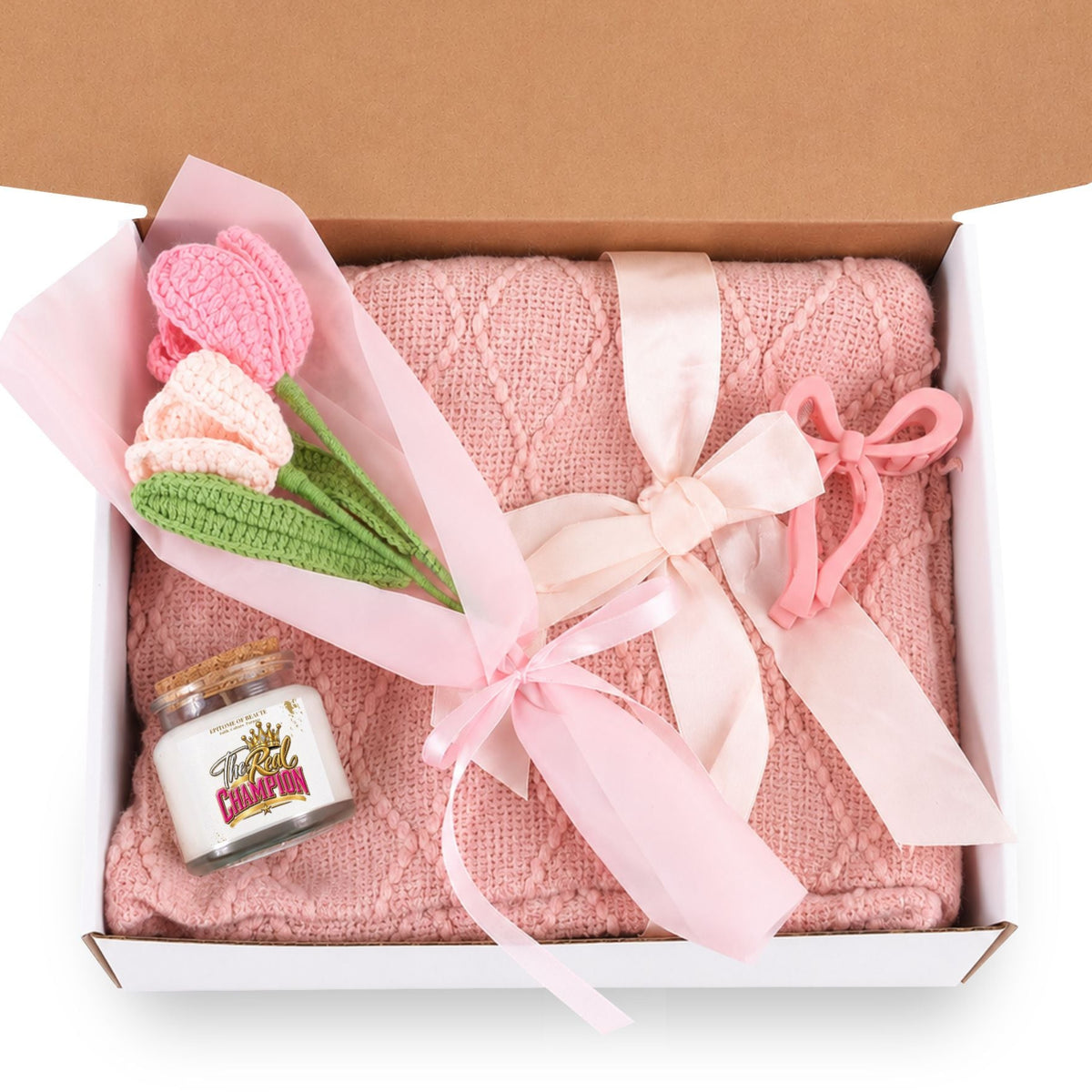 A pink knitted blanket or throw gift set presented in an open white box with a brown lid, tied with a pale pink ribbon. The set includes a small bouquet of crocheted pink and peach tulips with green stems and a small decorative candle jar with a gold elephant illustration.