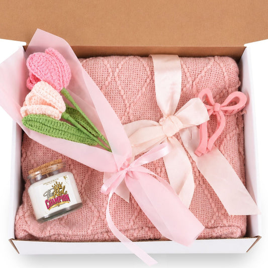 A pink knitted blanket or throw gift set presented in an open white box with a brown lid, tied with a pale pink ribbon. The set includes a small bouquet of crocheted pink and peach tulips with green stems and a small decorative candle jar with a gold elephant illustration.
