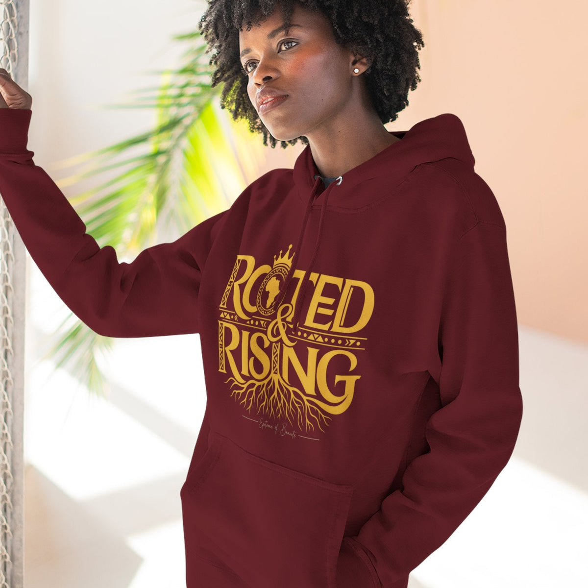 A woman wearing a maroon hoodie with a gold graphic that reads 'ROOTED & RISING' with roots and a crown, standing in a brightly lit indoor setting.