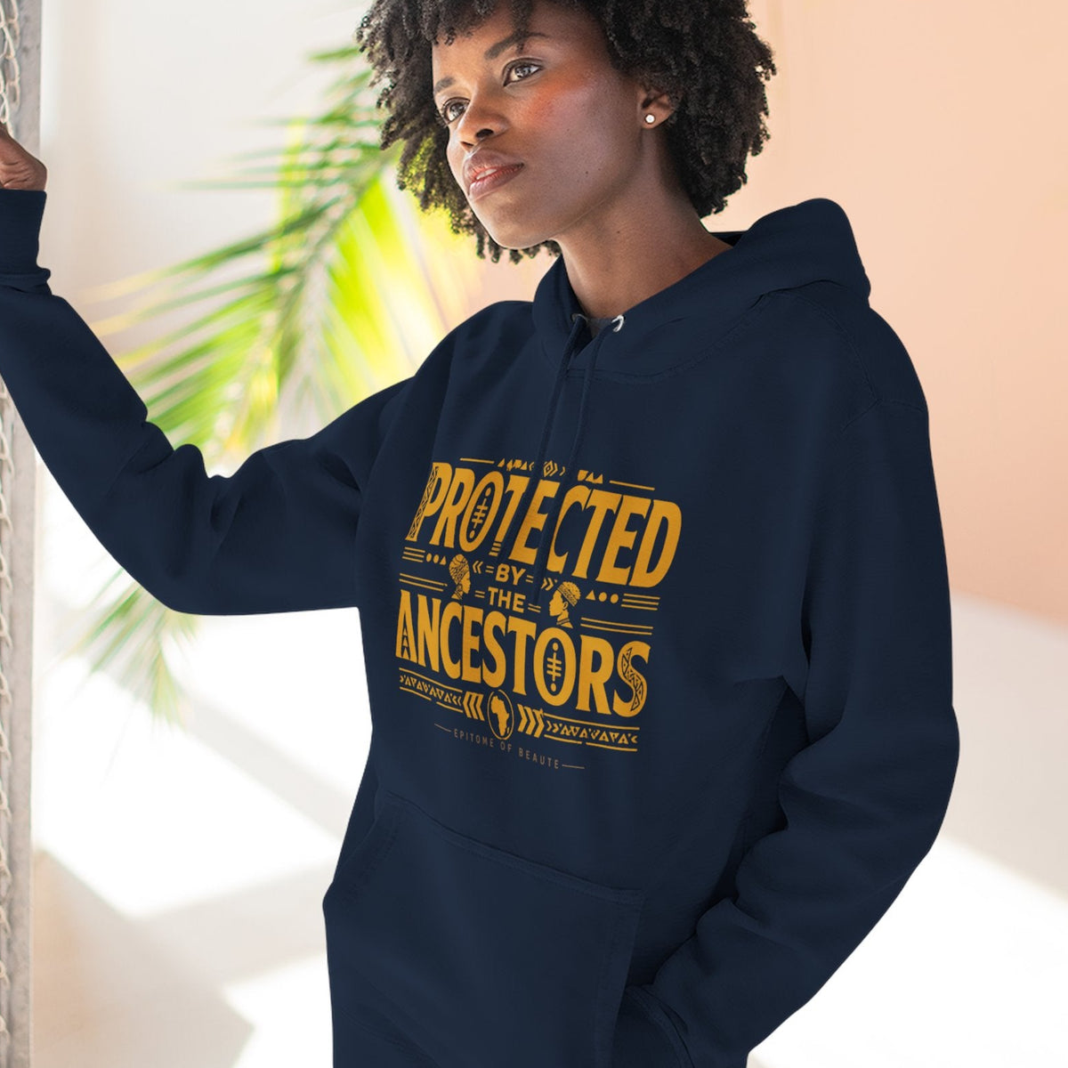 A woman wearing a navy blue hoodie with a yellow graphic print that reads 'PROTECTED BY THE ANCESTORS' stands next to a chain-link fence in a bright, sunlit indoor setting.