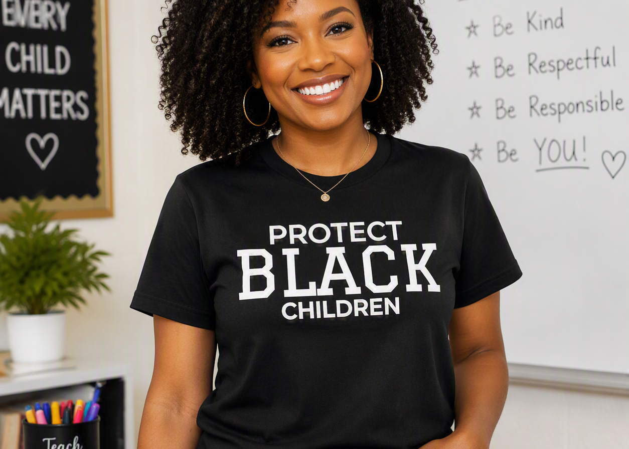 A smiling woman with curly hair stands in a classroom wearing a black graphic T-shirt with the text 'PROTECT BLACK CHILDREN' in white. The background includes a whiteboard with handwritten goals, a chalkboard, and a small plant.